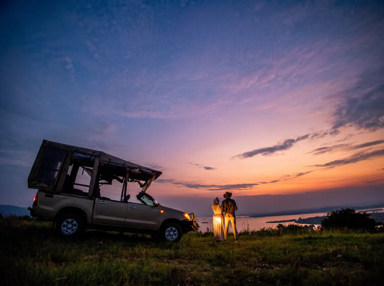 Guests beside a safari vehicle watching the sunset over the water.